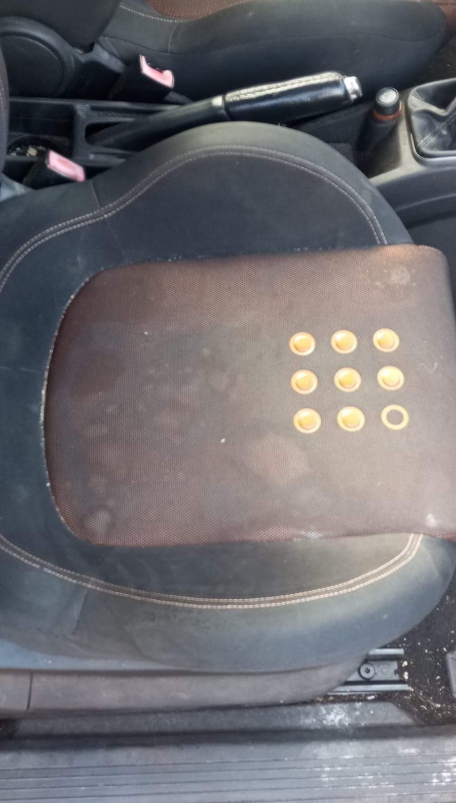 Brudne, czarne siedzenie samochodowe z brązową, siatkową nakładką z dziewięcioma okrągłymi wgłębieniami, z których jedno jest puste.