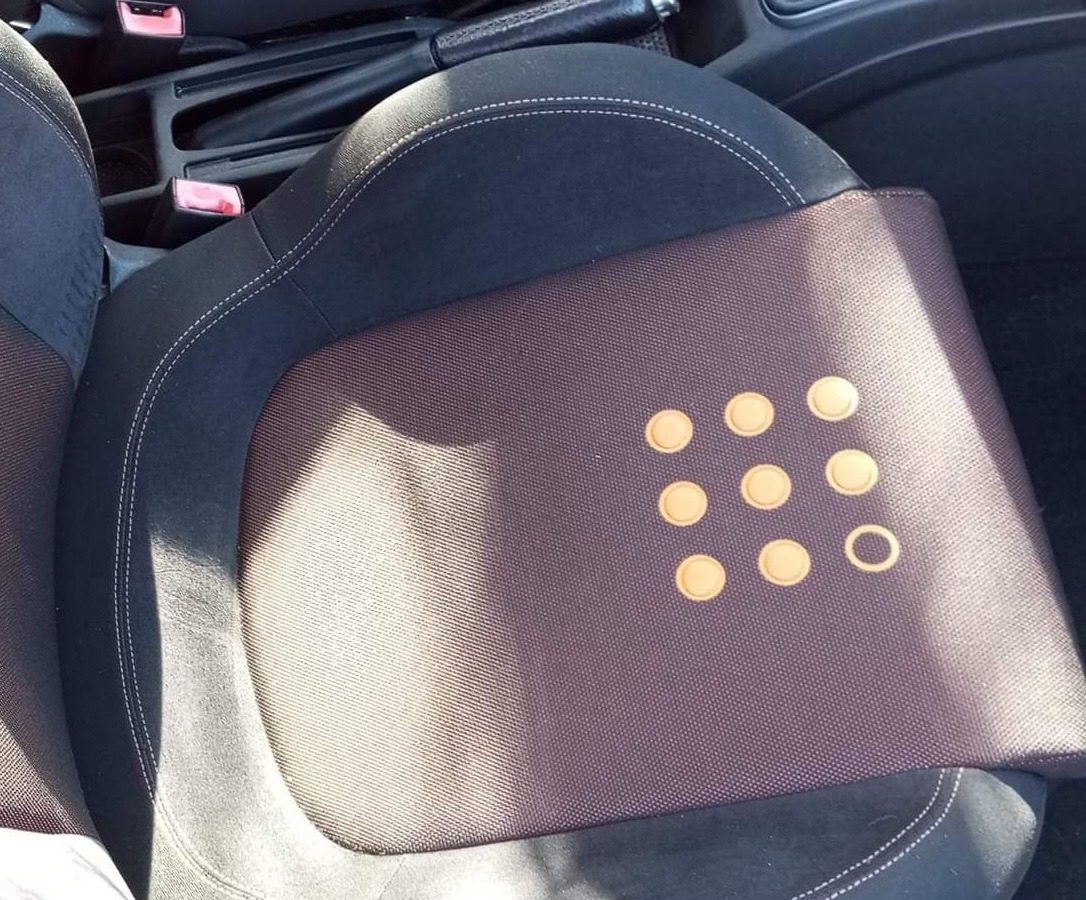 Fotel samochodowy z brązową nakładką z dziewięcioma okrągłymi wgłębieniami, jedno z nich puste, widoczne szwy i fragment wnętrza pojazdu.