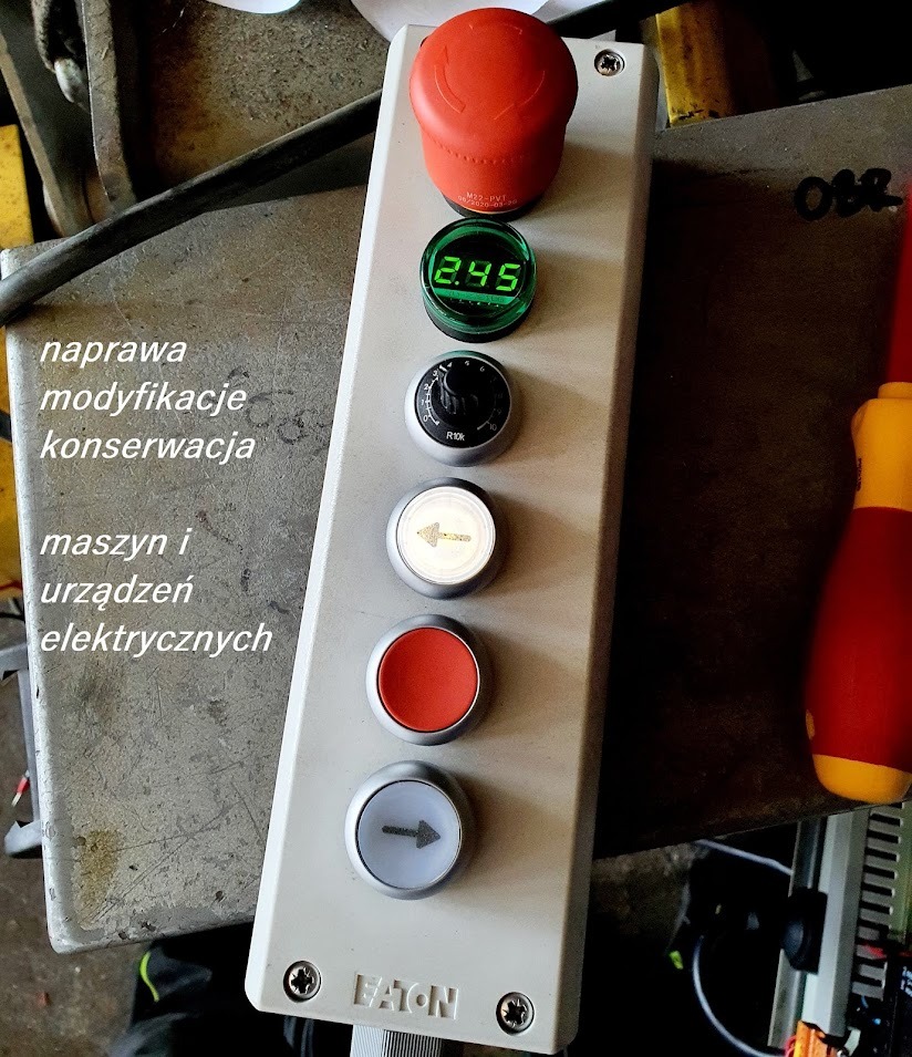 Panel sterowania urządzeniem elektrycznym z przyciskami, w tym czerwonym przyciskiem awaryjnym, wyświetlaczem cyfrowym i pokrętłem, umieszczony na metalowej powierzchni roboczej.