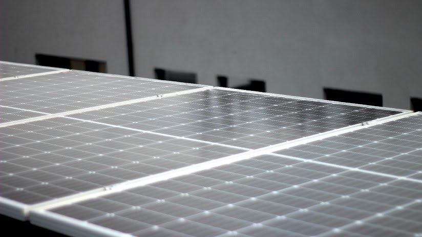 Rząd paneli słonecznych na dachu budynku, widoczne moduły fotowoltaiczne o strukturze krzemowej, szare tło ściany z cieniami.