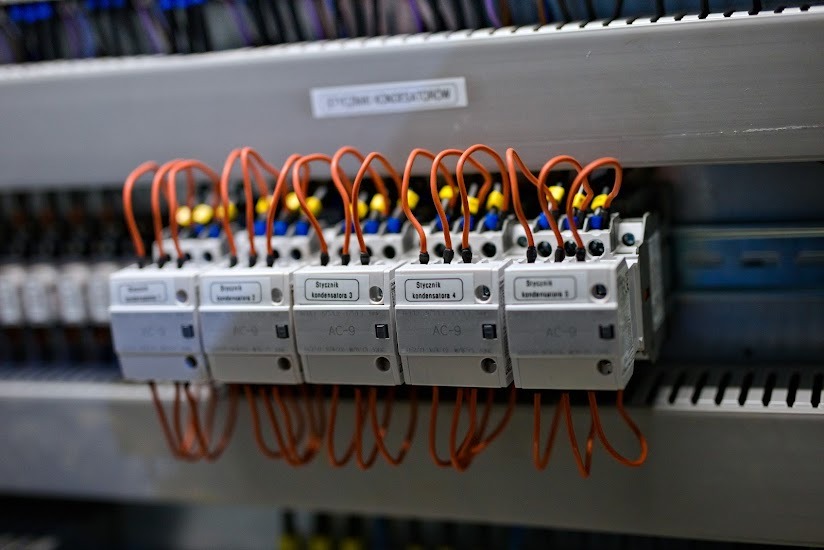 Szereg styczników kondensatorów AC-9 z pomarańczowymi przewodami podłączonych do szyny montażowej w szafie sterowniczej.