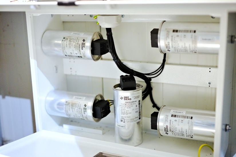 Wnętrze białej szafy sterowniczej z zamontowanymi cylindrycznymi kondensatorami firmy ZEZ SILKO, połączonymi czarnymi przewodami. Widoczne etykiety i oznaczenia na obudowach kondensatorów.