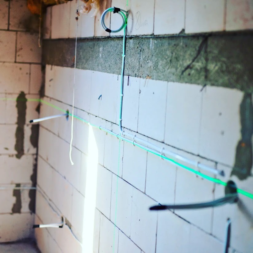 Ściana z betonu komórkowego z poziomymi liniami lasera wyznaczającymi trasy kabli elektrycznych, pionowy kabel przytwierdzony do ściany uchwytami.