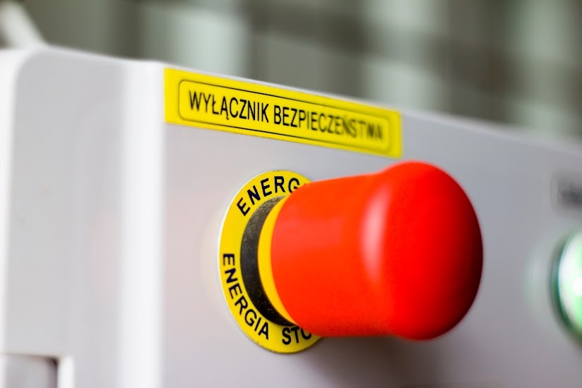 Czerwony przycisk bezpieczeństwa z żółtą obwódką i napisem 'Energia Stop' na szarej obudowie, obok żółta naklejka z napisem 'Wyłącznik Bezpieczeństwa'.