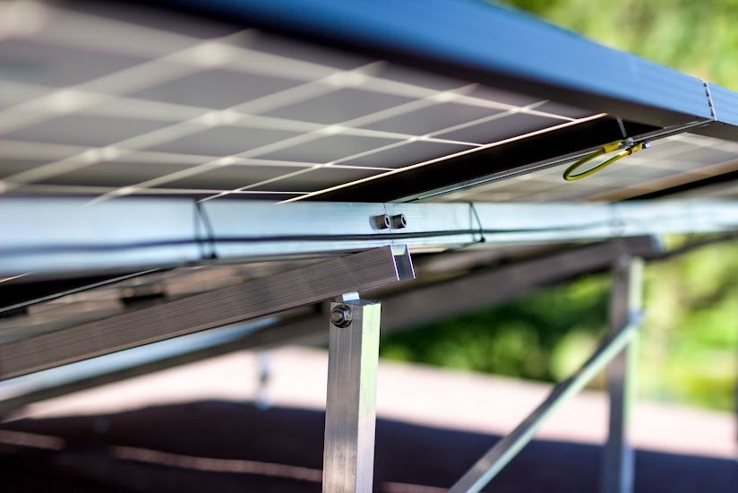 Fragment konstrukcji wsporczej paneli fotowoltaicznych z widocznymi elementami mocującymi, uziemieniem i profilem aluminiowym na tle rozmytej zieleni.