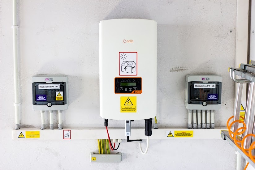 Biały inwerter fotowoltaiczny Solis zamontowany na ścianie, z podłączonymi przewodami i dwoma szarymi rozdzielnicami PV-AC i PV-DC po bokach, widoczne żółte oznaczenia ostrzegawcze.