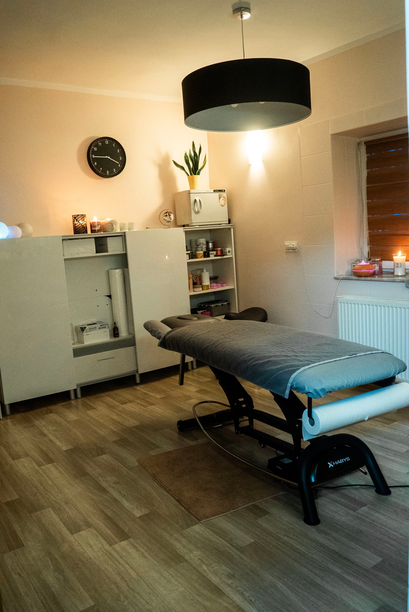 Profesjonalny gabinet z elektrycznie regulowanym stołem do zabiegów, pokrytym szarym prześcieradłem i rolką papieru, oświetlony wiszącą lampą, z szafkami i zegarem na ścianie.