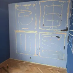 Niebieski pokój w trakcie przygotowania do prac wykończeniowych, widoczne ślady po usuniętych elementach dekoracyjnych na ścianie, obrysowane żółtą taśmą malarską, parkiet w jodełkę, narzędzia...