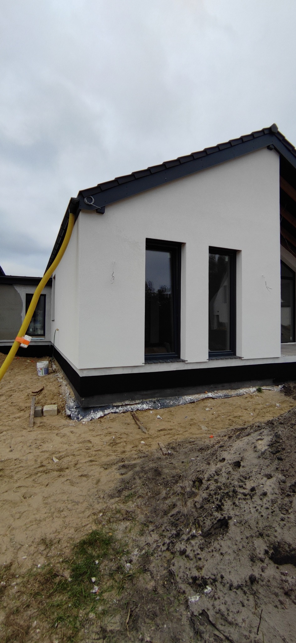 Świeżo otynkowana, biała elewacja domu jednorodzinnego z czarnymi ramami okiennymi, widoczna żółta rura wentylacyjna i przygotowany teren wokół budynku.