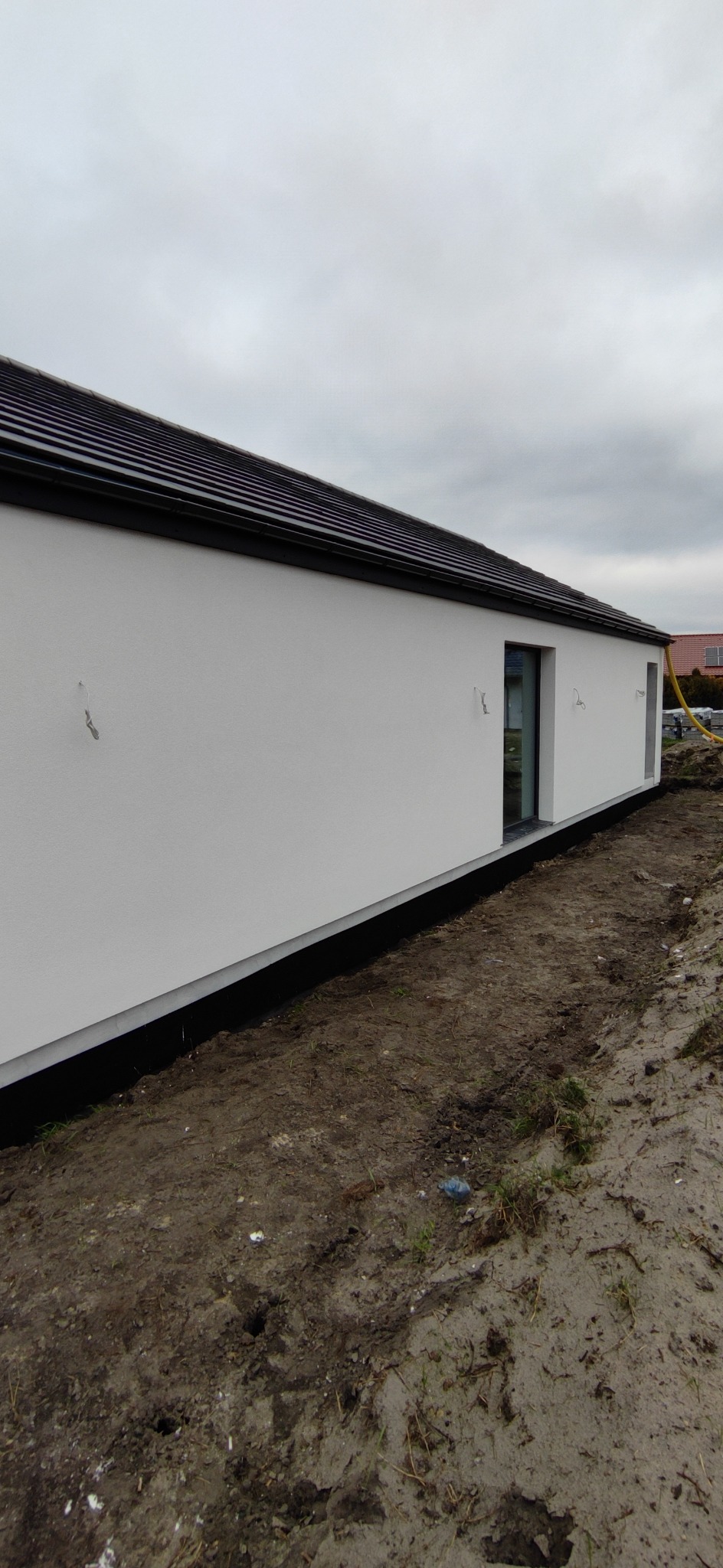 Boczny widok na świeżo otynkowany, biały dom jednorodzinny z czarnym dachem i obramowaniem okien, na tle pochmurnego nieba i przygotowanego terenu.