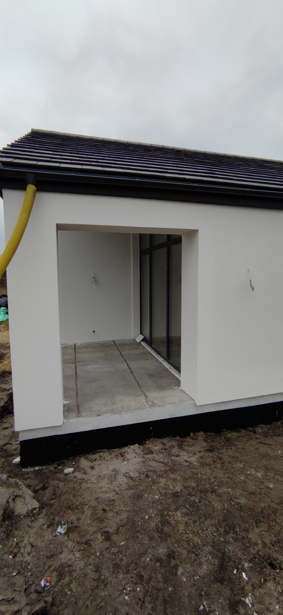 Nowo otynkowana i pomalowana elewacja budynku z widocznym fragmentem dachu, orynnowaniem i surowym betonem w otworze drzwiowym.