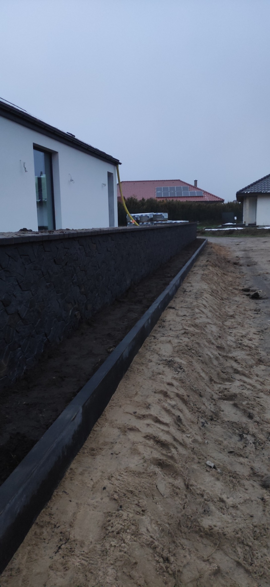 Nowo wybudowany murek z kamienia elewacyjnego, oddzielający teren posesji od drogi, z widocznymi elementami domu i sąsiednich budynków w tle.