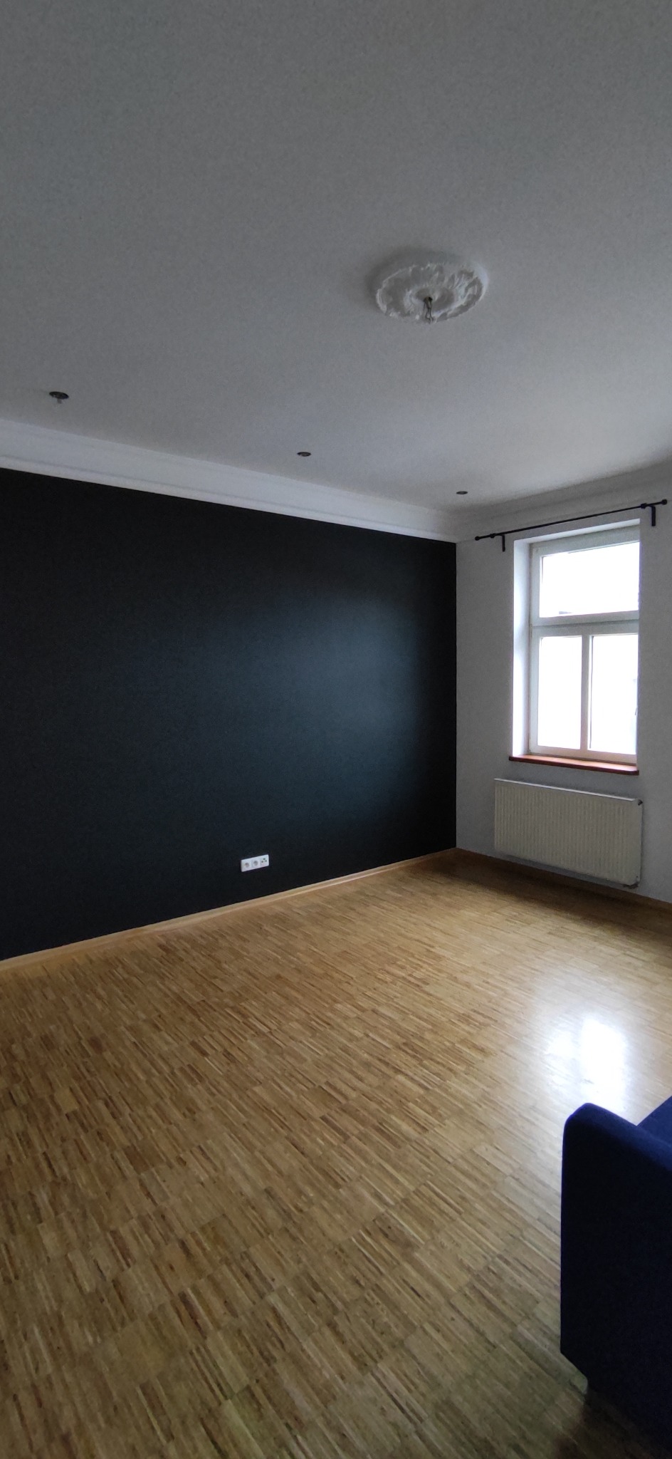 Pomalowany na czarno fragment ściany w pustym pokoju z parkietem, biały sufit z ozdobną rozetą, grzejnik pod oknem.