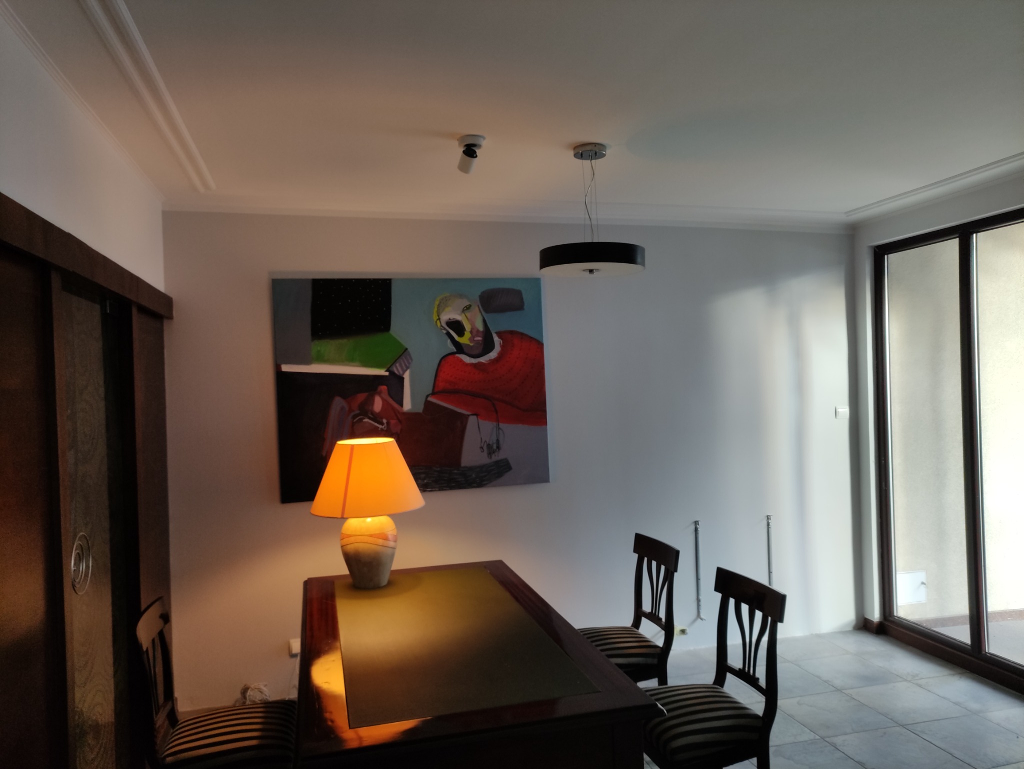 Jasne wnętrze pokoju z biurkiem oświetlonym lampką, dwoma krzesłami w paski, obrazem na ścianie i dużym oknem balkonowym.