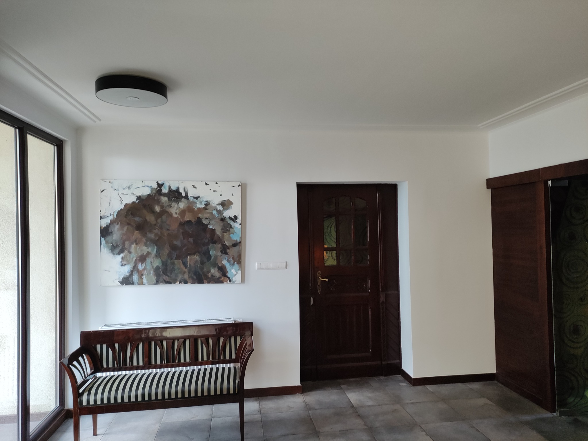 Jasne wnętrze z białymi ścianami, ciemnymi drewnianymi drzwiami z szybkami, ławką w czarno-białe pasy i abstrakcyjnym obrazem na ścianie. Na suficie okrągła lampa.