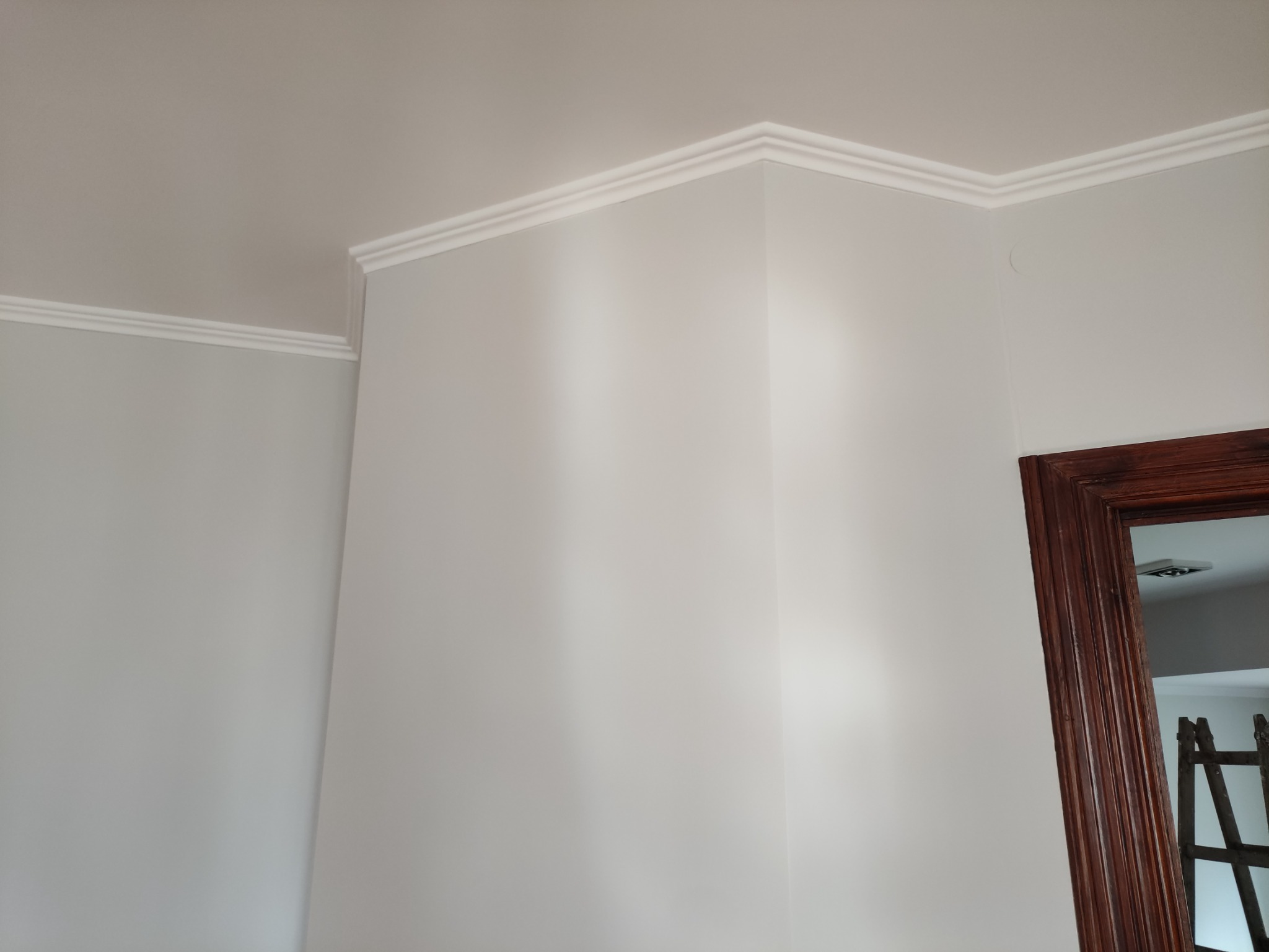 Świeżo pomalowany narożnik pokoju z delikatną listwą dekoracyjną pod sufitem, widoczna część futryny drzwi i drabina w tle.