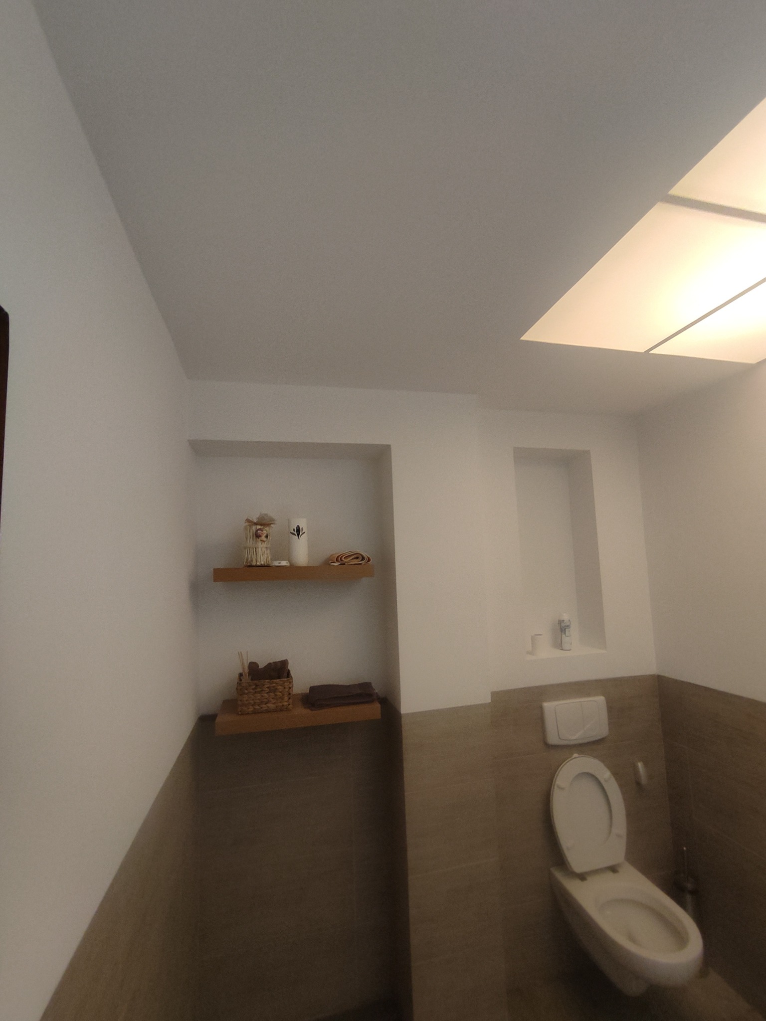 Jasna łazienka z białymi ścianami, drewnianymi półkami z dekoracjami i beżowymi płytkami, oświetlona panelami LED w suficie, z widoczną toaletą.