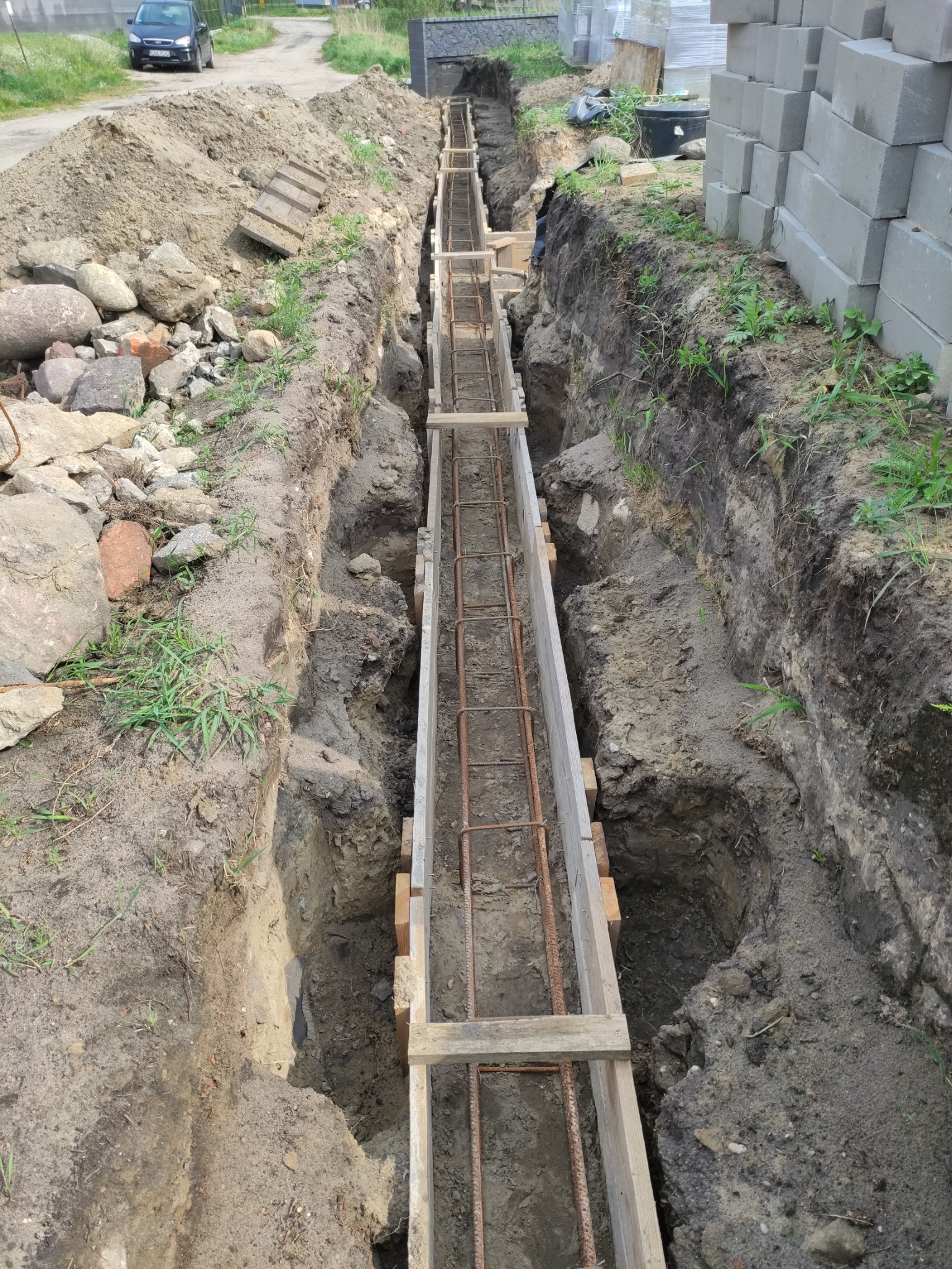 Przygotowanie fundamentów: wykop z drewnianym szalunkiem i zbrojeniem stalowym, widok z góry, obok sterta bloczków betonowych.