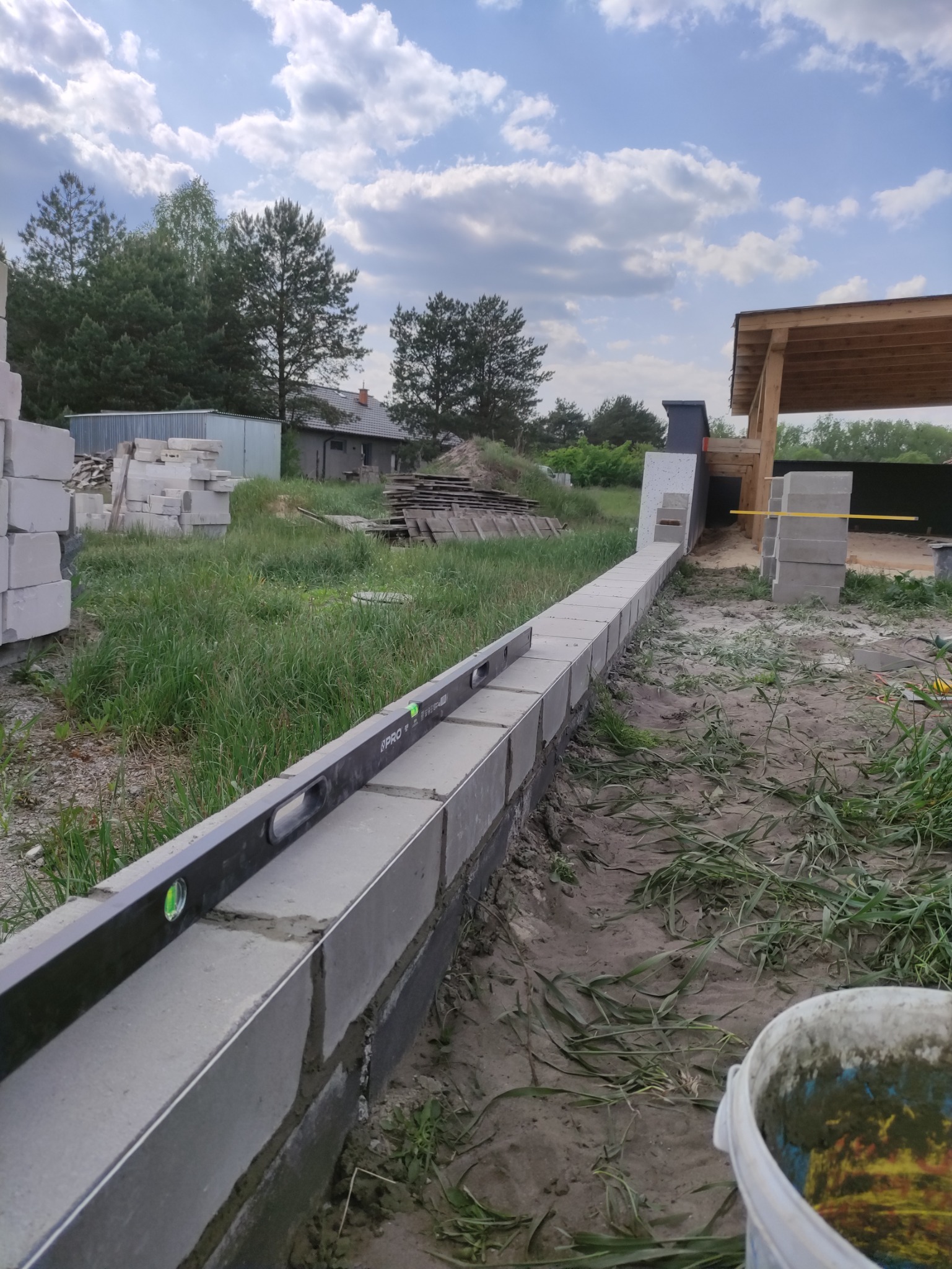 Wyrównywanie bloczków betonowych poziomnicą podczas budowy fundamentów domu jednorodzinnego, widoczny fragment konstrukcji drewnianej zadaszenia i stosy bloczków w tle.