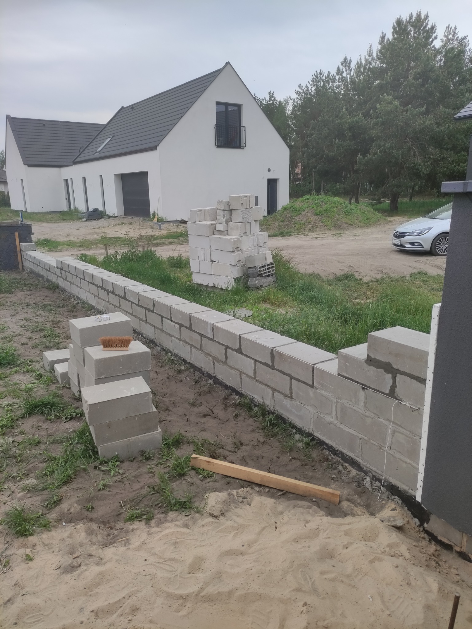 Budowa ogrodzenia z bloczków betonowych na tle nowego domu z szarą dachówką i białymi ścianami, widoczne narzędzia i materiały budowlane.