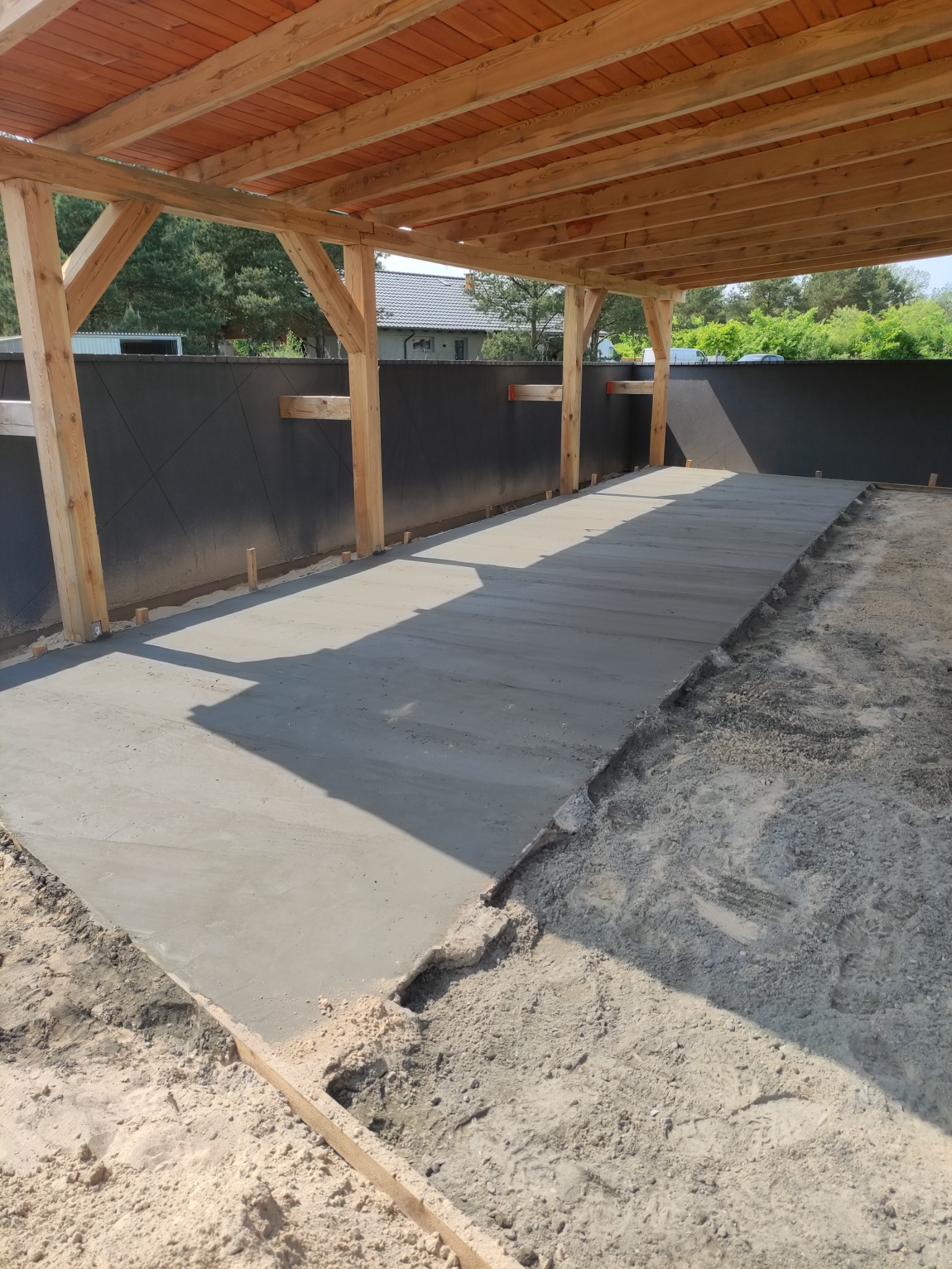 Świeżo wylana betonowa posadzka pod zadaszeniem z drewnianą konstrukcją, widoczne szalunki i przygotowane podłoże.