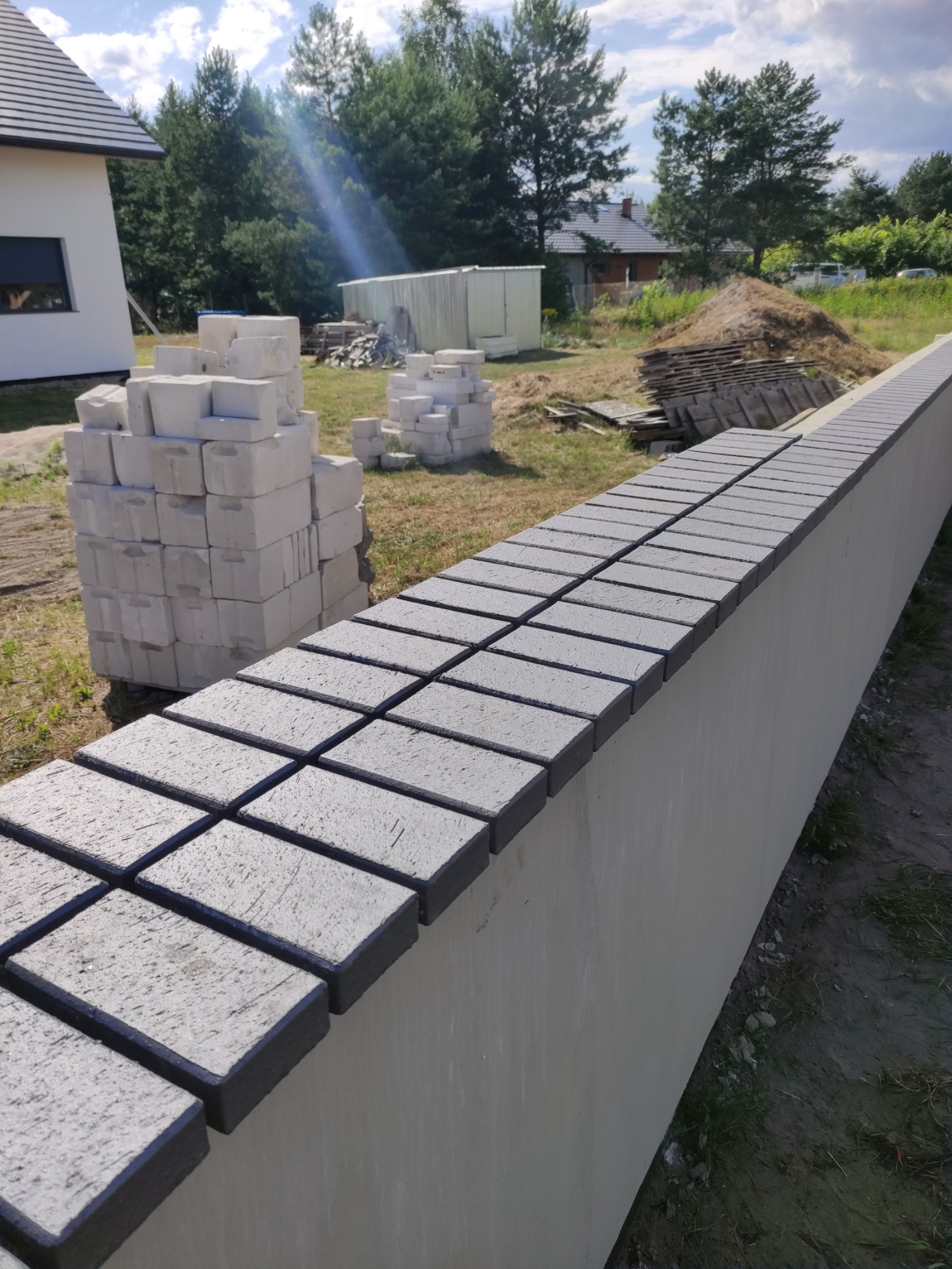 Niski murek ogrodzeniowy z szarymi, betonowymi płytkami na górze, w tle dom jednorodzinny i sterta materiałów budowlanych.