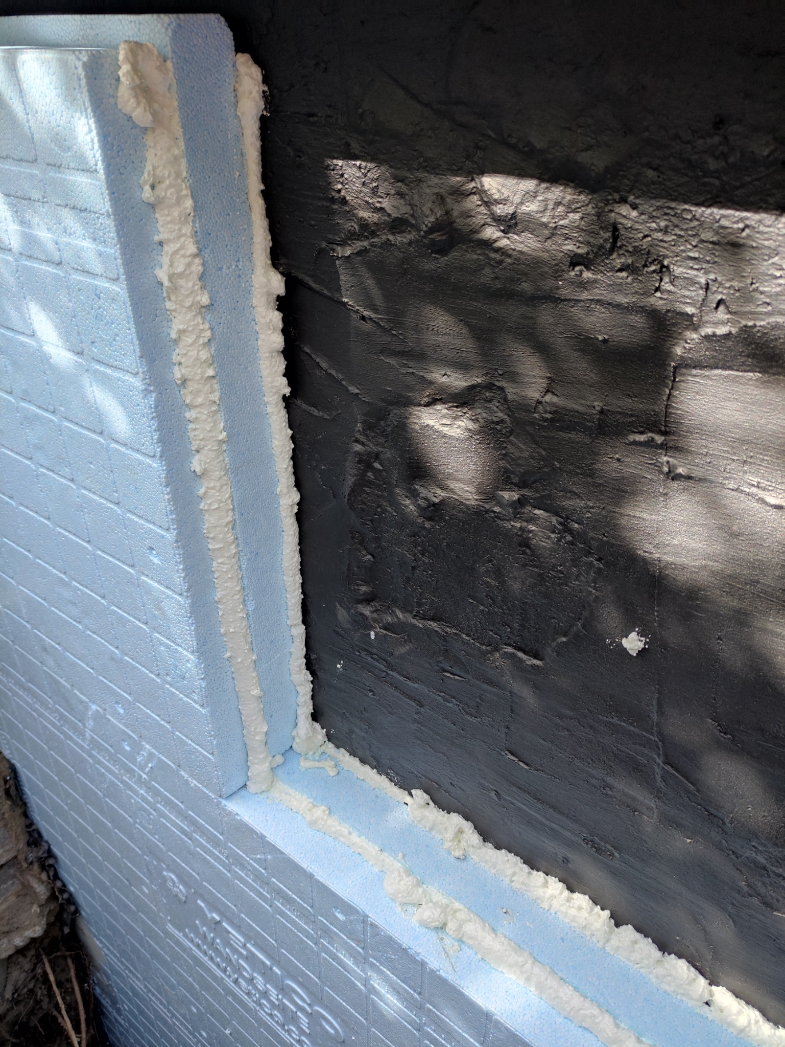 Narożnik budynku z nałożonymi niebieskimi płytami styropianowymi, przymocowanymi pianką montażową do ciemnej, chropowatej elewacji w trakcie prac ociepleniowych.