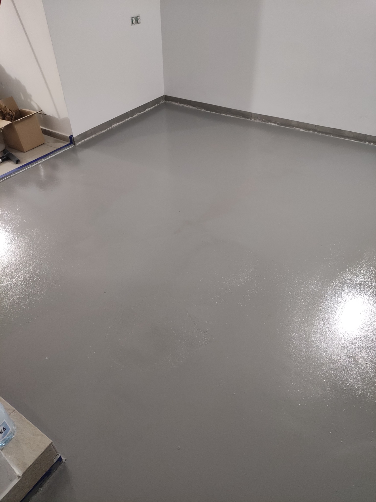 Świeżo wylana, gładka, szara posadzka żywiczna w pomieszczeniu z białymi ścianami i listwami przypodłogowymi, widoczny karton i butelka wody w rogu.