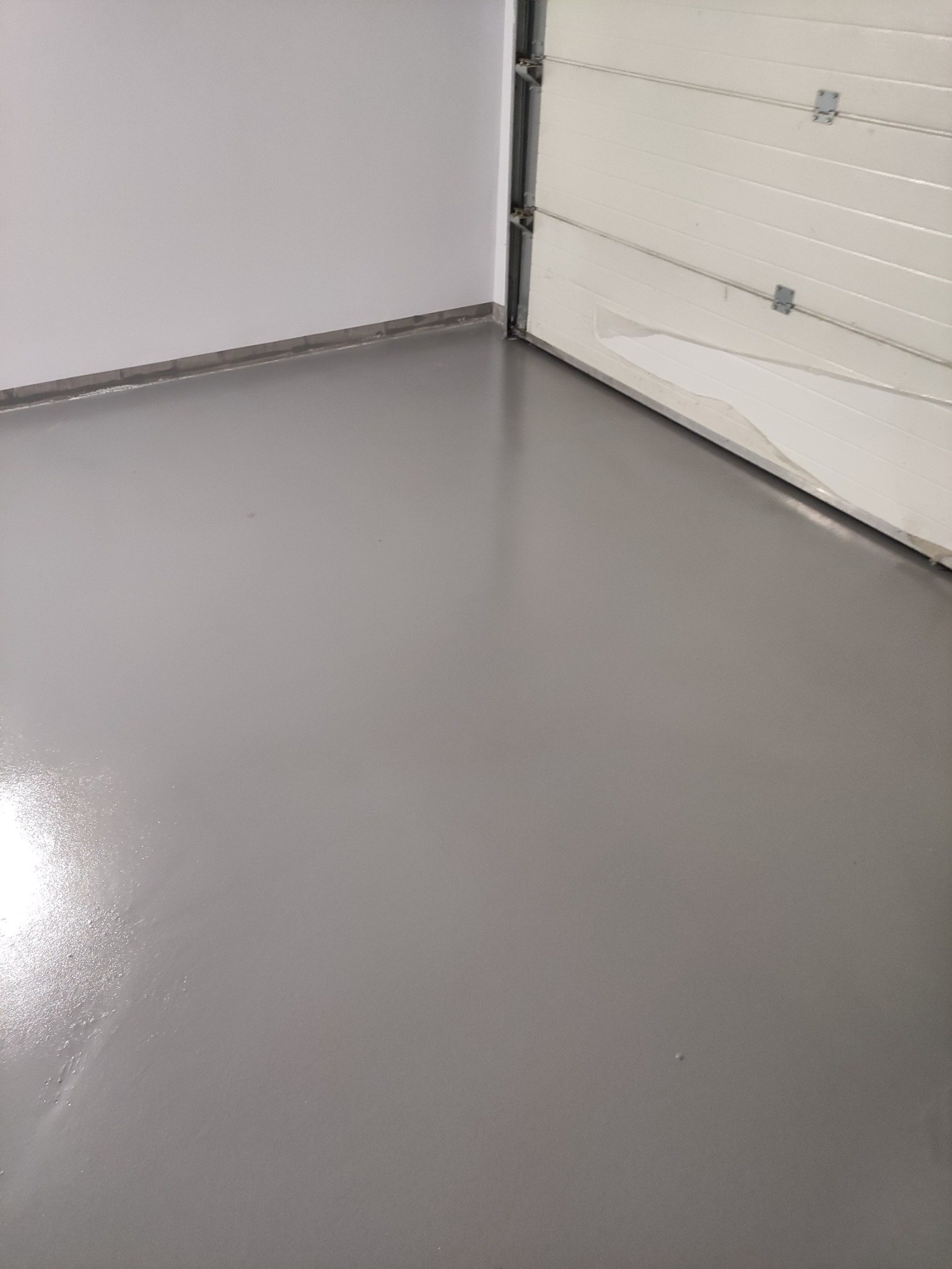 Świeżo wylana, szara posadzka żywiczna w garażu, z częściowo otwartą białą bramą segmentową i pomalowaną na biało ścianą.