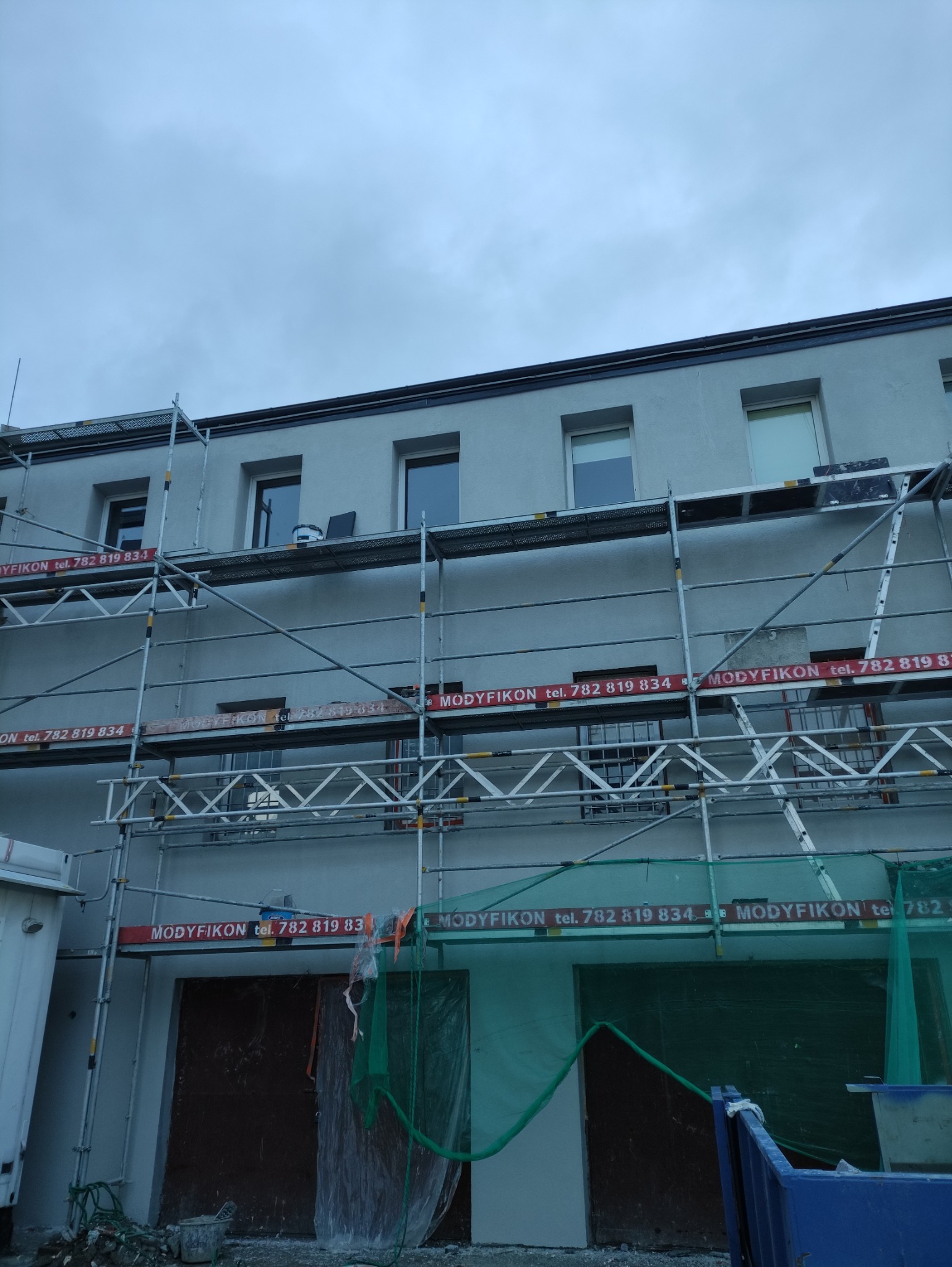 Elewacja budynku w trakcie malowania, widoczne rusztowania z reklamami firmy MODYFIKON, ślady farby i pojemnik na rusztowaniu.