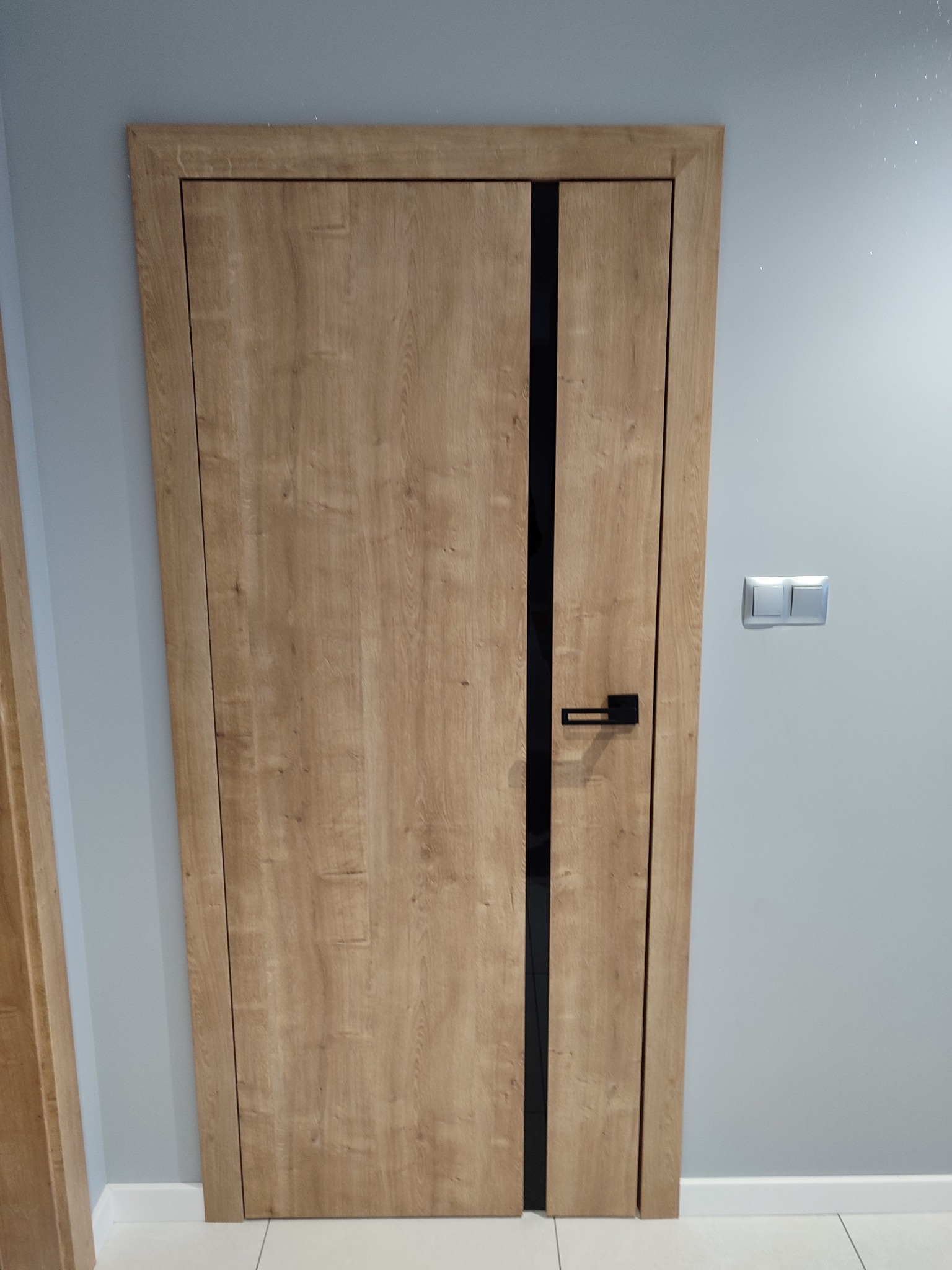 Drewniane drzwi wewnętrzne z czarnym pionowym akcentem i klamką, zamontowane w szarej ścianie z białymi listwami przypodłogowymi i włącznikiem światła.