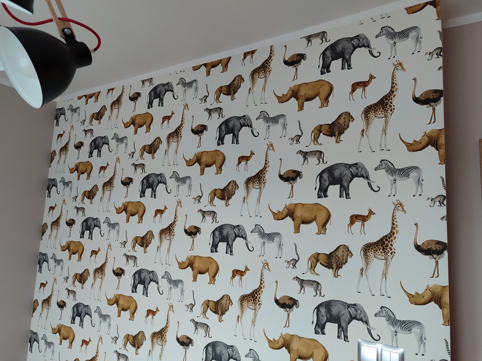 Ściana pokryta tapetą z motywem afrykańskich zwierząt, takich jak żyrafy, słonie, zebry i lwy, oświetlona lampą.