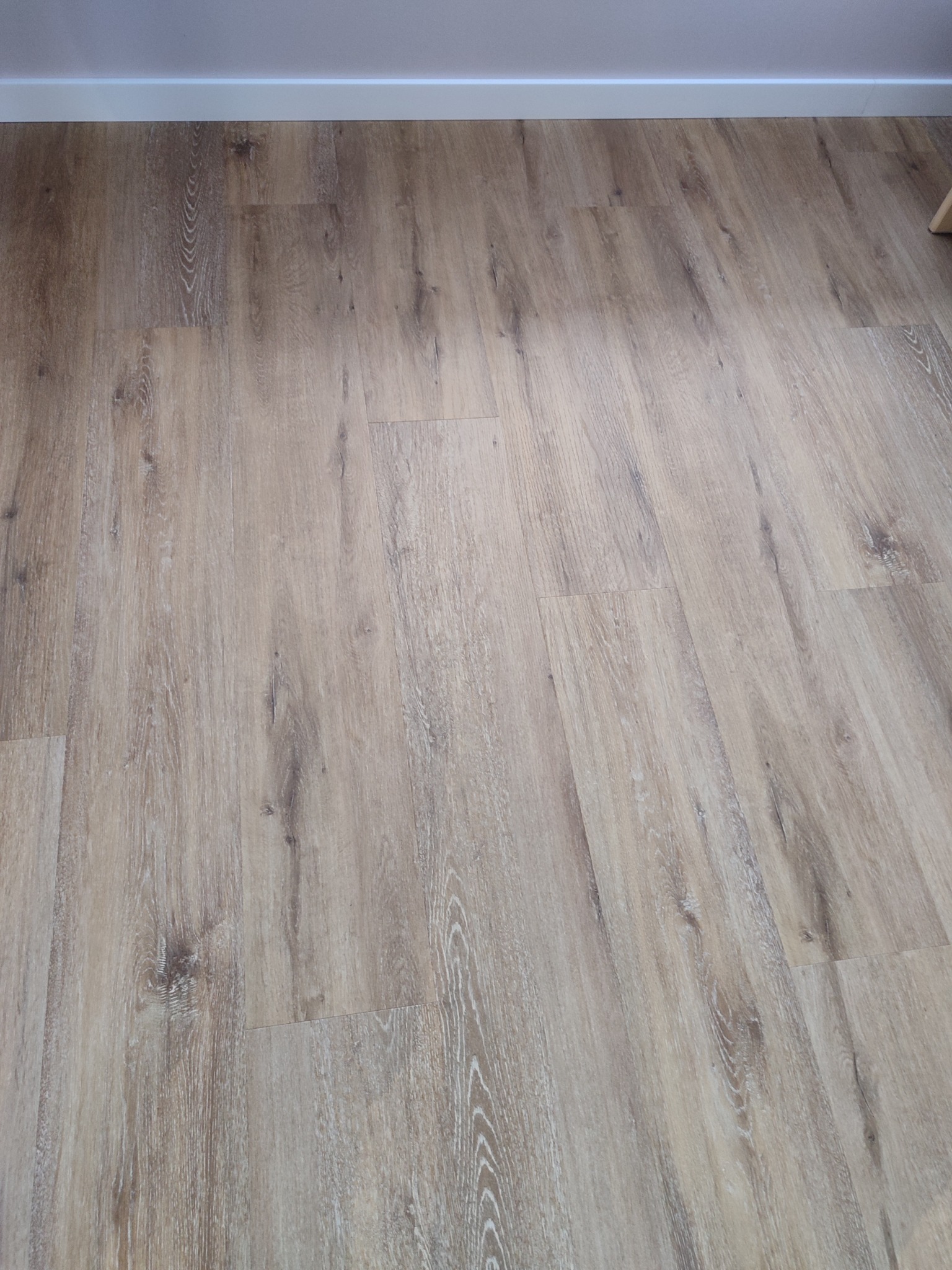 Jasnobrązowe panele podłogowe z wyraźną strukturą drewna, ułożone równolegle do ściany z białą listwą przypodłogową.