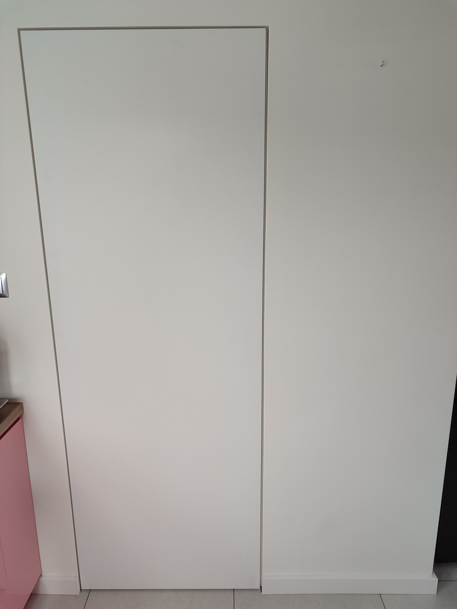 Białe, ukryte drzwi w ścianie, z widoczną minimalistyczną listwą przypodłogową i fragmentem różowej szafki po lewej stronie.
