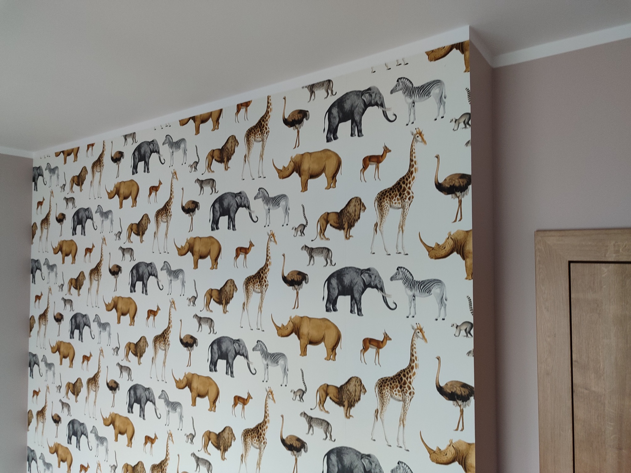 Ściana pokryta tapetą z motywem afrykańskich zwierząt: słonie, żyrafy, zebry, lwy, strusie, nosorożce i antylopy, widoczna przy białym suficie i różowej ścianie.