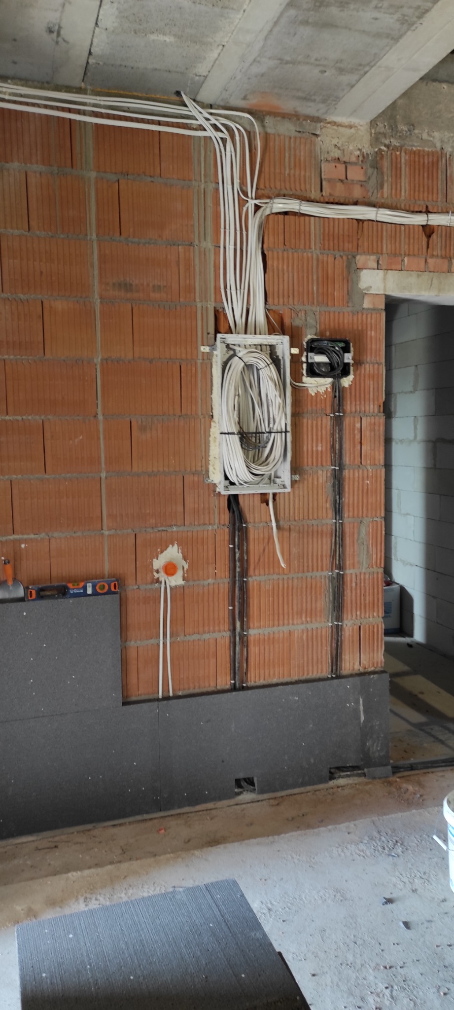 Instalacja elektryczna w trakcie montażu w budynku w stanie surowym: przewody ułożone w korytkach i puszkach na murze z cegły, częściowo zakrytym szarą płytą izolacyjną.