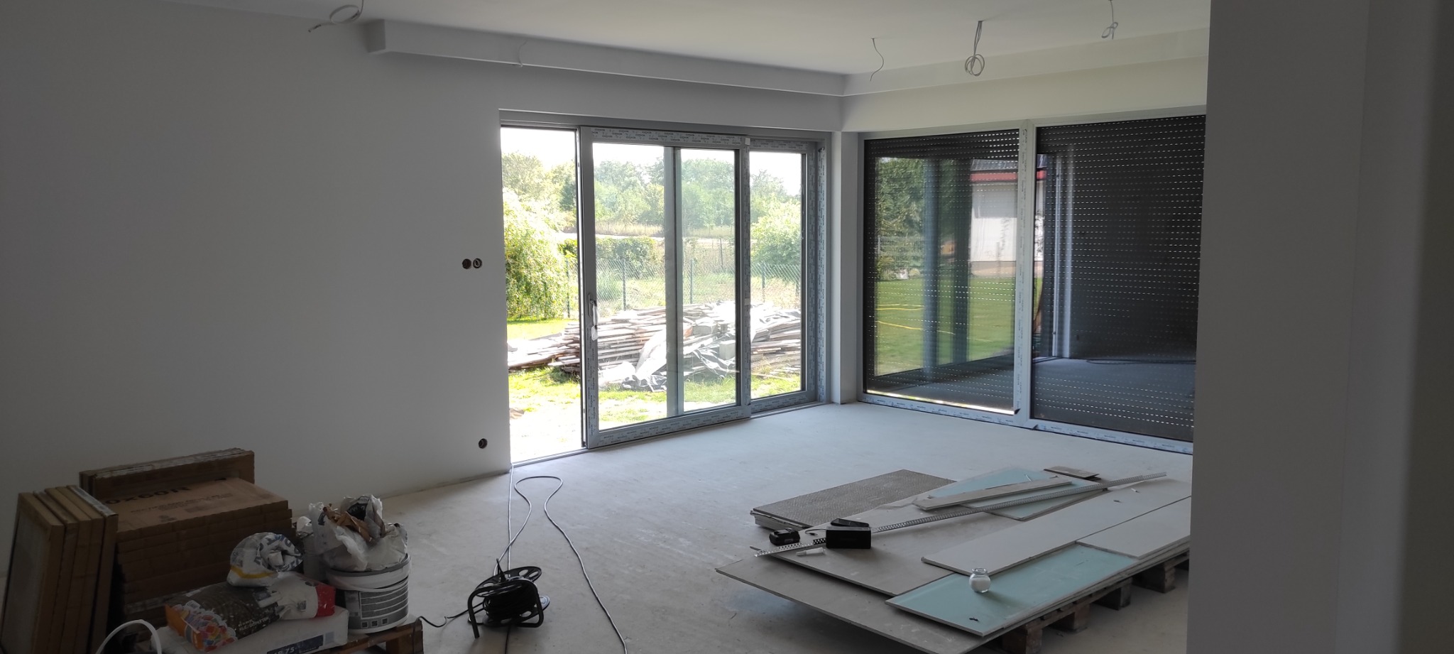 Wnętrze w trakcie prac wykończeniowych: przygotowane ściany, duże okna przesuwne z widokiem na ogród, paleta z płytami gipsowo-kartonowymi i narzędziami, puszki z farbą i wiadro z zaprawą.