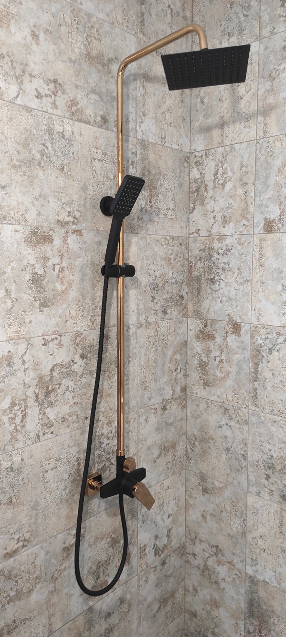 Nowoczesny zestaw prysznicowy: czarna deszczownica i słuchawka na miedzianej rurze, zamontowany na ścianie wyłożonej płytkami w odcieniach beżu i szarości.