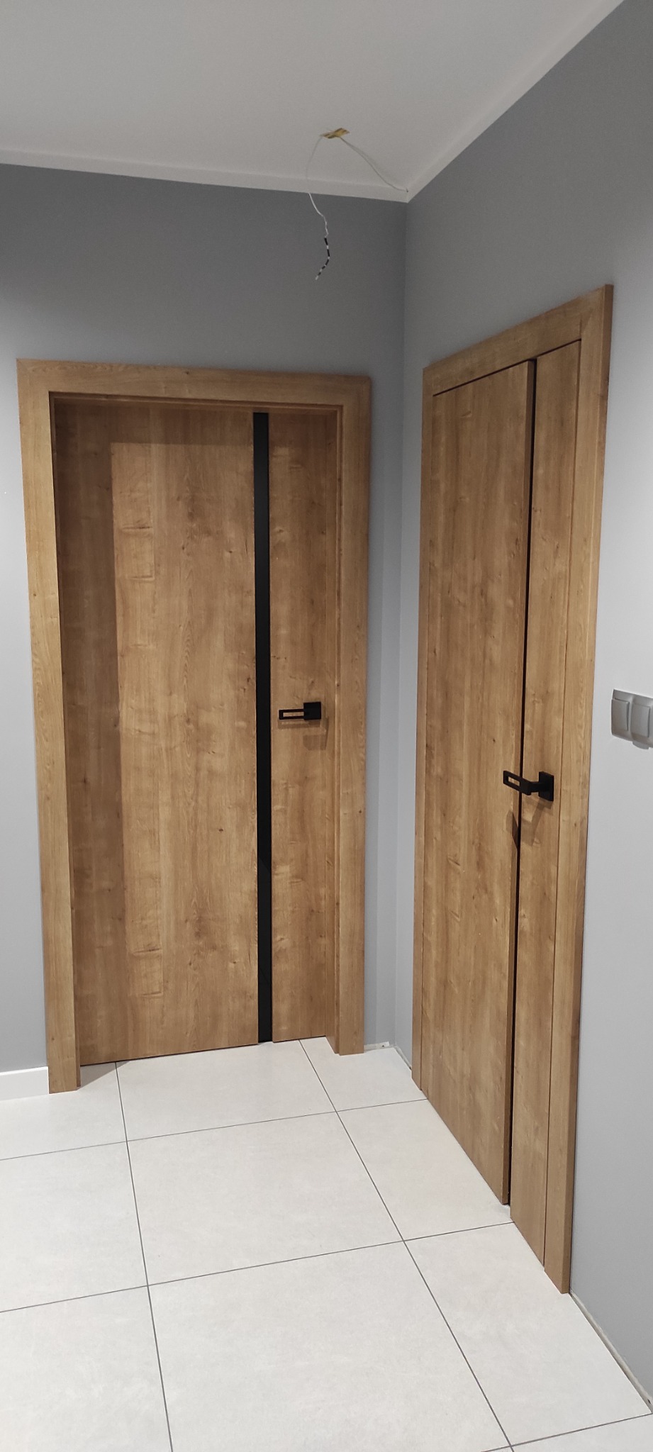 Dwa zamontowane, drewnopodobne drzwi wewnętrzne z czarnymi klamkami i pionowym czarnym paskiem, na białej podłodze z płytek, szare ściany, wystające kable z sufitu.