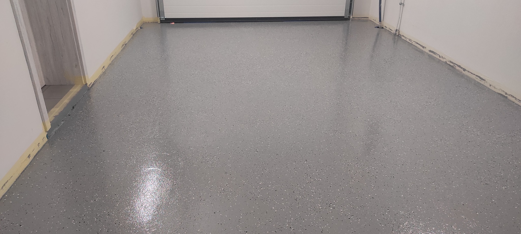 Świeżo wylana, szara posadzka żywiczna z drobnymi, ciemnymi plamkami w garażu, zabezpieczona taśmą malarską przy ścianach i bramie garażowej.