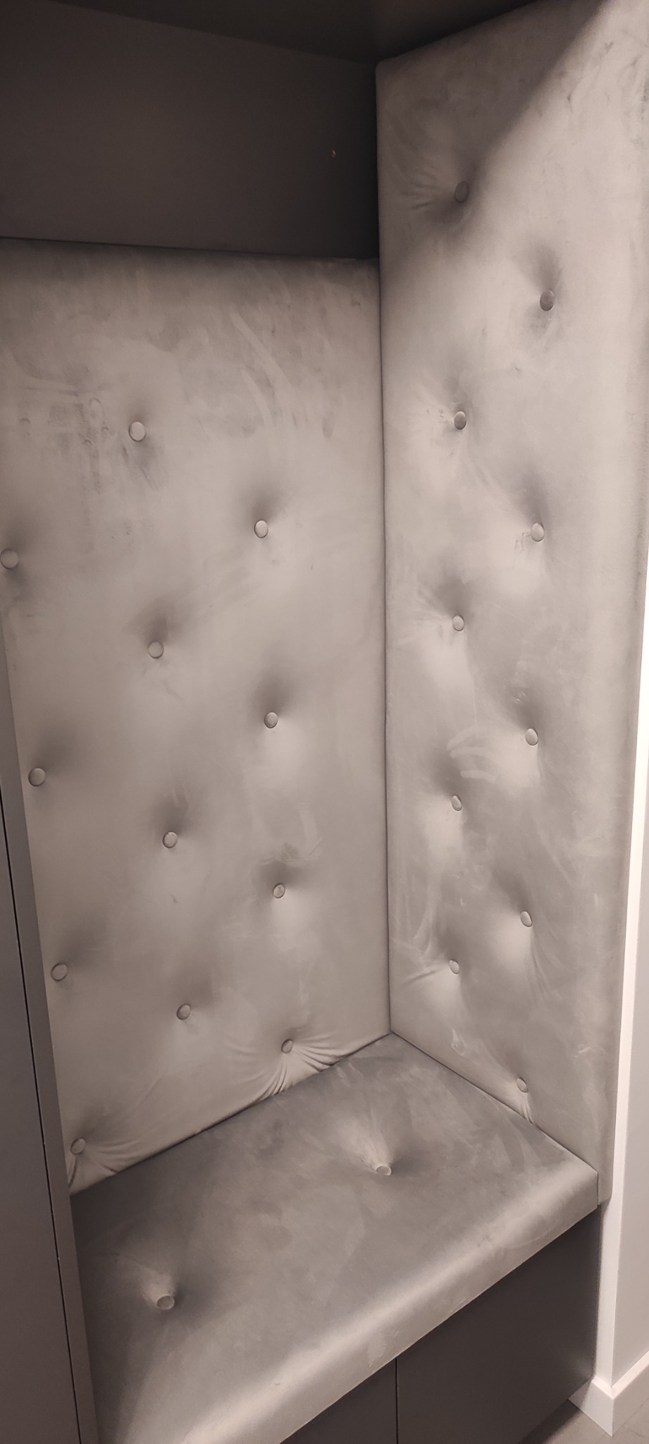 Tapicerowany narożnik z siedziskiem i oparciem w kolorze szarym, wykończony dekoracyjnymi guzikami, wnęka z zabudową meblową.