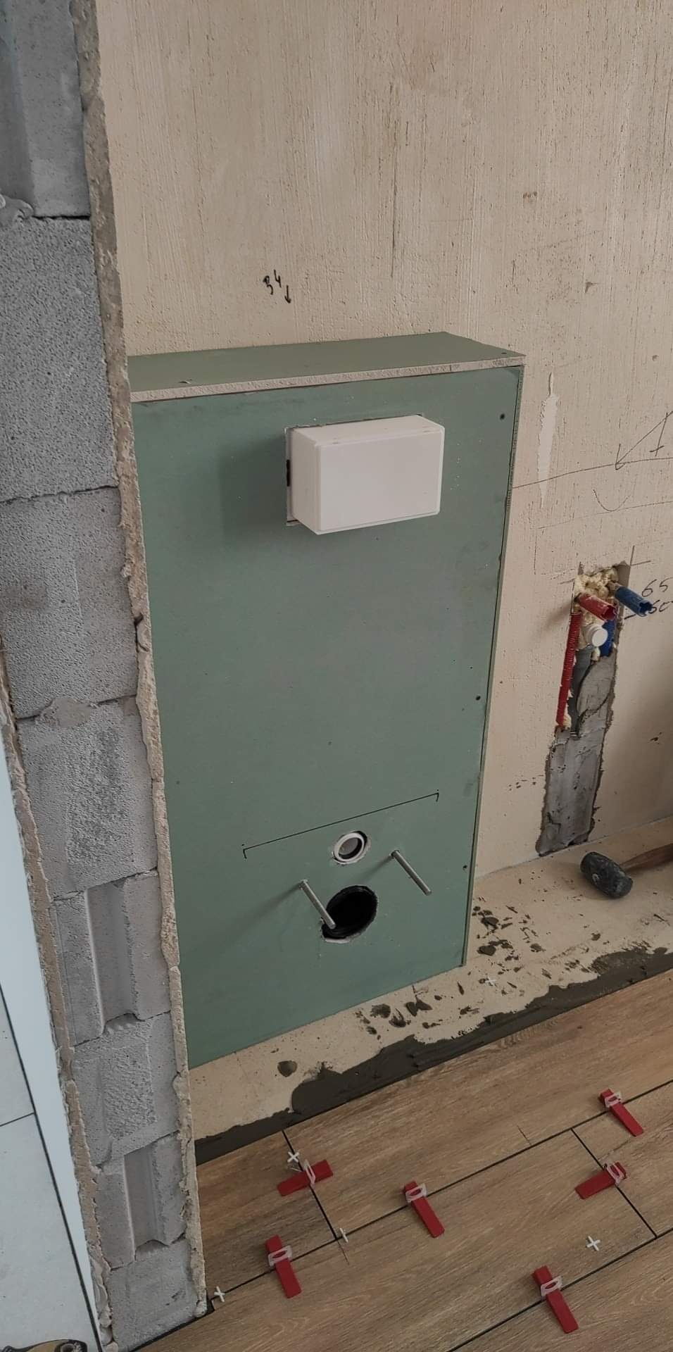 Montaż stelaża podtynkowego z płyt karton gips w trakcie wykańczania łazienki, widoczne płytki podłogowe z czerwonymi klipsami poziomującymi i instalacja wodna w ścianie.