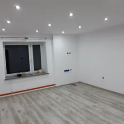 Pusty, nowo wykończony pokój z jasnymi panelami podłogowymi, białymi ścianami, oknem i punktowym oświetleniem sufitowym, widoczne przygotowane gniazdka elektryczne i rury grzewcze w kolorze...