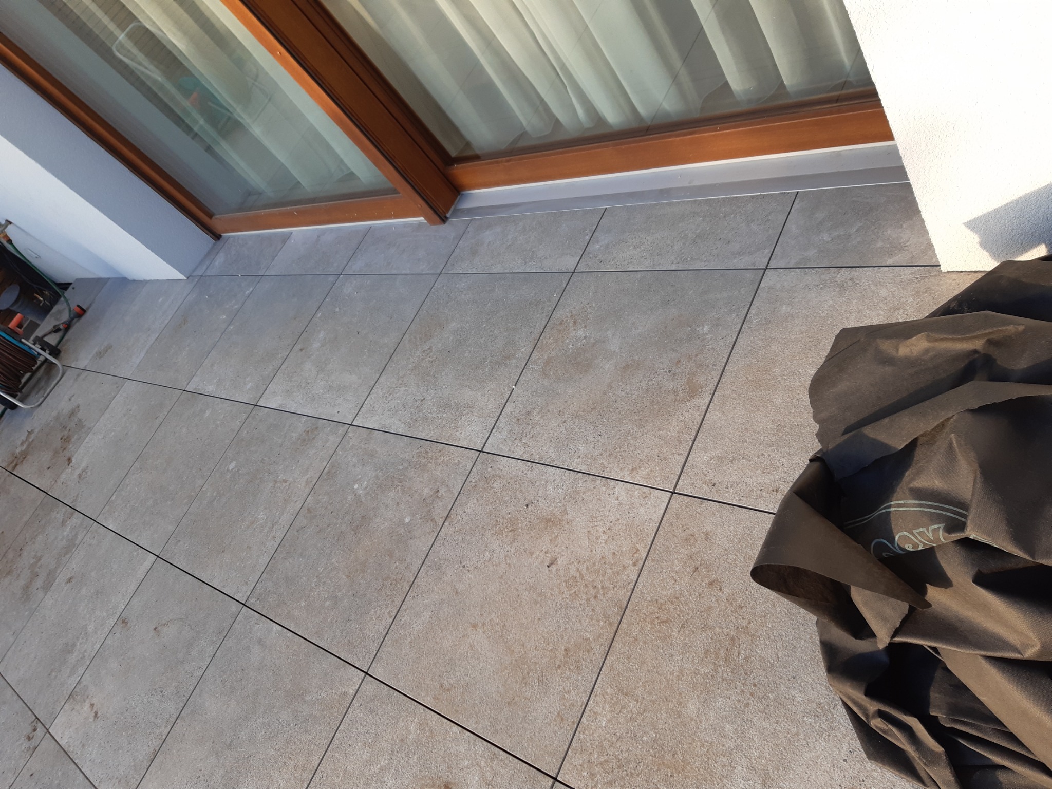 Wyłożony jasnymi płytkami taras z widocznym oknem balkonowym i fragmentem czarnej plandeki ochronnej.