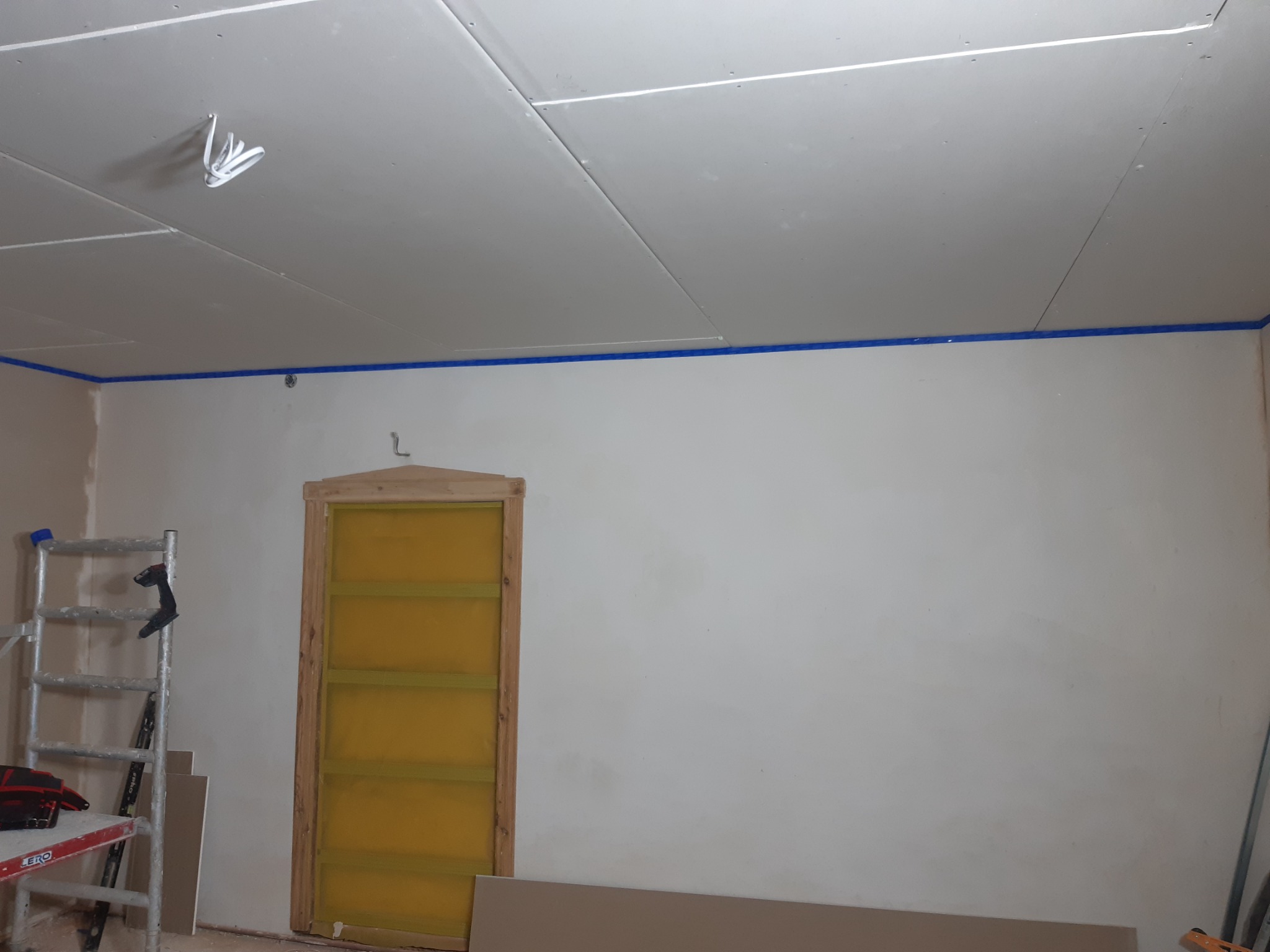 Pokój w trakcie remontu: zamontowane płyty karton gips na suficie, zabezpieczone taśmą malarską ściany, drabina i narzędzia w rogu, surowe obramowanie drzwi.