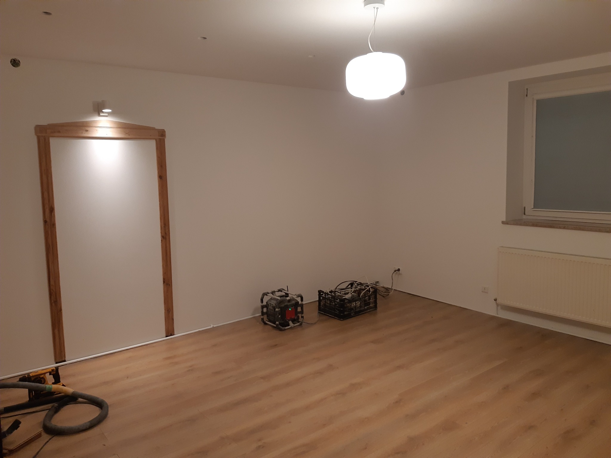 Pokój w trakcie wykańczania: drewniana podłoga, białe ściany z zamontowanym ościeżem drzwiowym, grzejnik i lampa sufitowa; widoczne narzędzia do obróbki podłogi i oświetlenie budowlane.