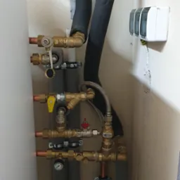 Szczegółowe ujęcie instalacji hydraulicznej z zaworami, rurami miedzianymi i izolacją termiczną, widoczny manometr i skrzynka elektryczna na ścianie.