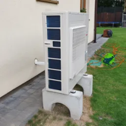 Zewnętrzna jednostka pompy ciepła zamontowana na betonowych podstawach przy ścianie budynku z widocznym przewodem w ścianie, na trawniku zwinięty wąż ogrodowy.