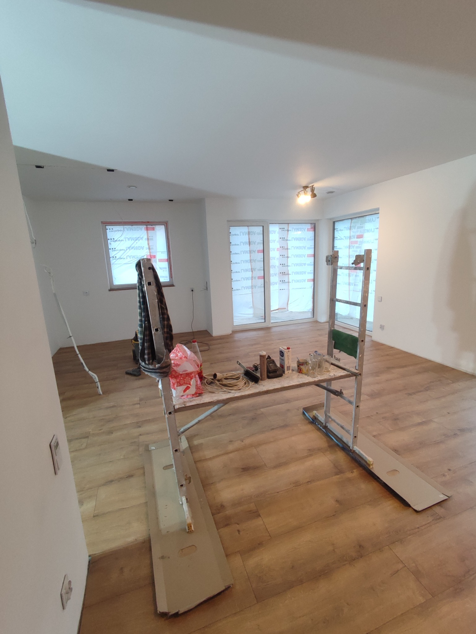 Wnętrze w trakcie przygotowań do prac wykończeniowych, z zabezpieczonymi oknami, rozstawionym rusztowaniem, drabiną i narzędziami na blacie roboczym.