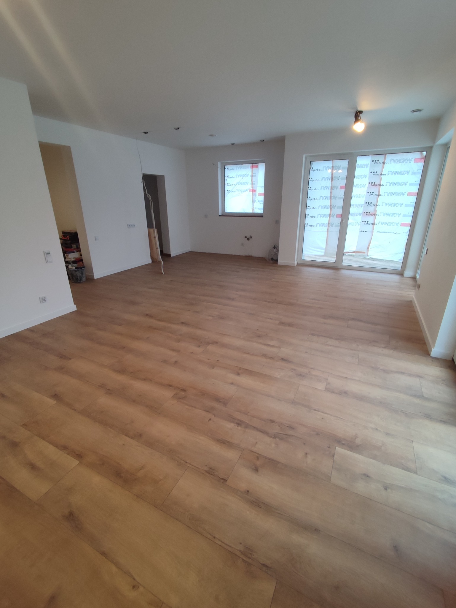 Jasne panele podłogowe ułożone w nowym, przestronnym wnętrzu z białymi ścianami i oknami zabezpieczonymi folią ochronną, przygotowane do dalszych prac wykończeniowych.